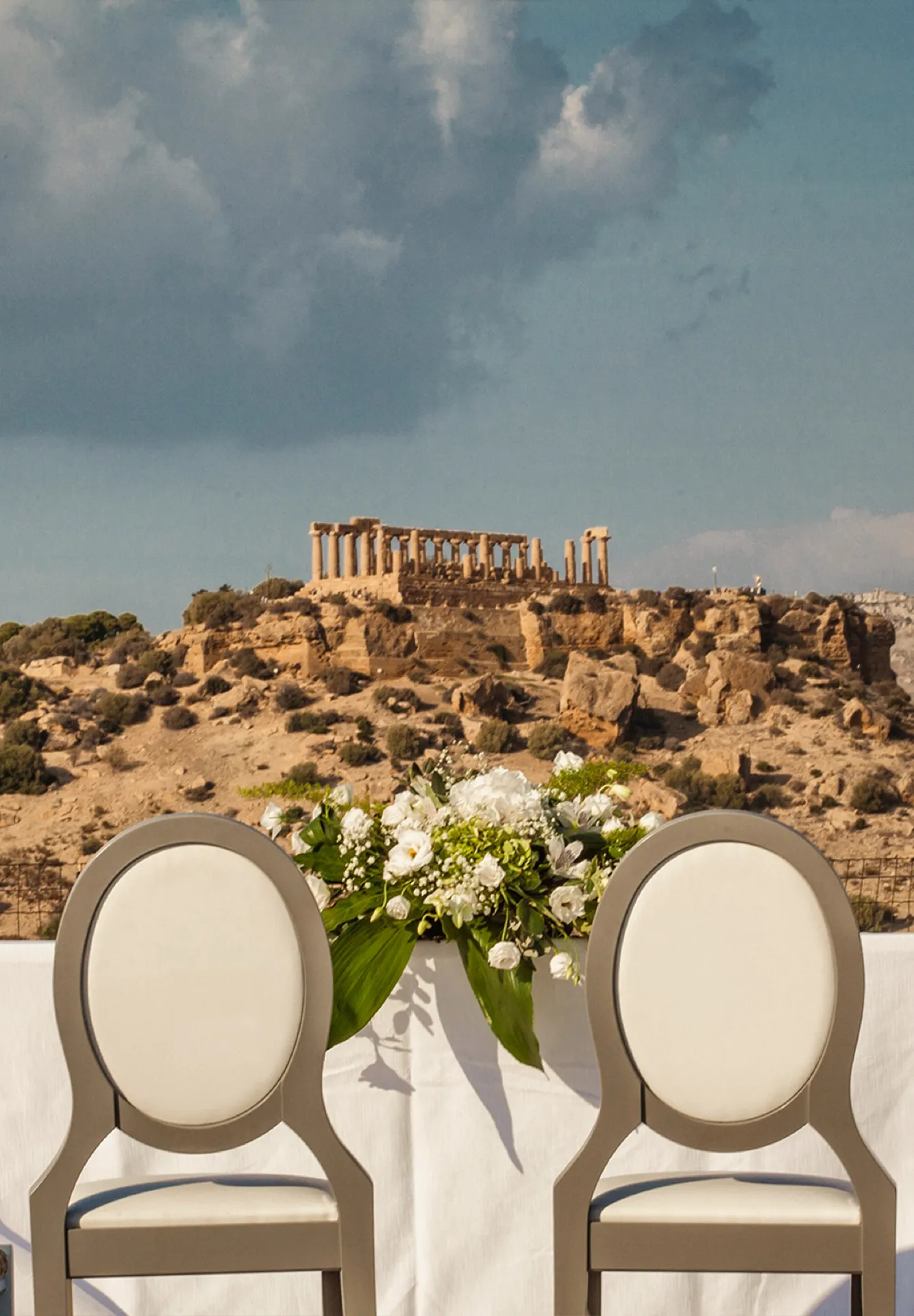 Allestimento per cerimonia nuziale con sedie, composizioni floreali bianche e tavolo decorato, con panorama sul tempio della Valle dei Templi sullo sfondo.
