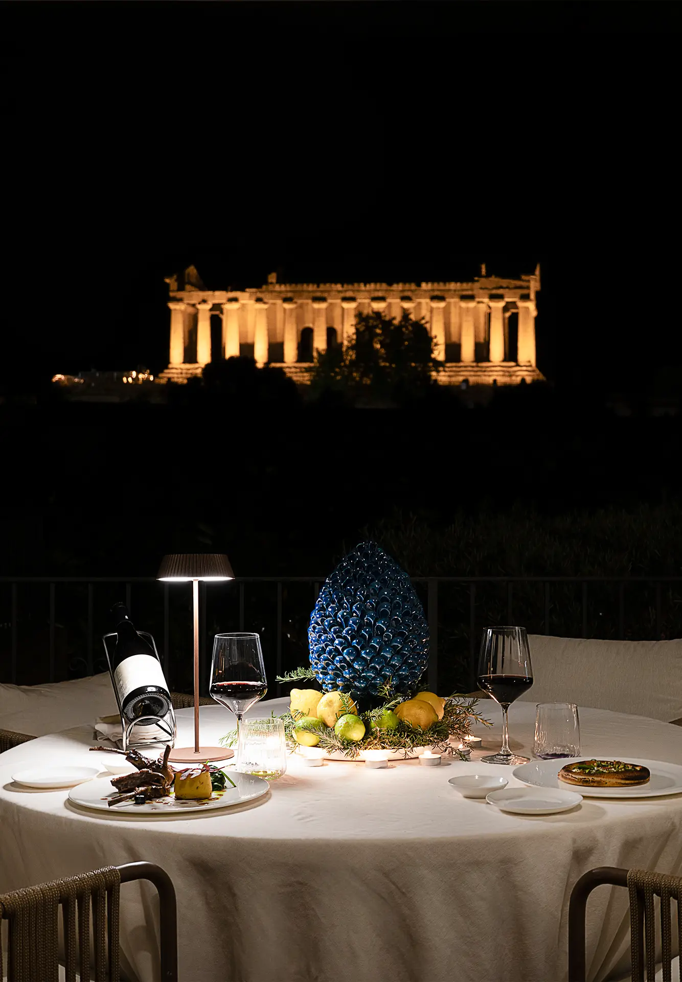 Tavolo del Ristorante Terrazza degli Dei apparecchiato di notte con vista sul tempio illuminato, vino, piatti gourmet e pigna in ceramica blu.