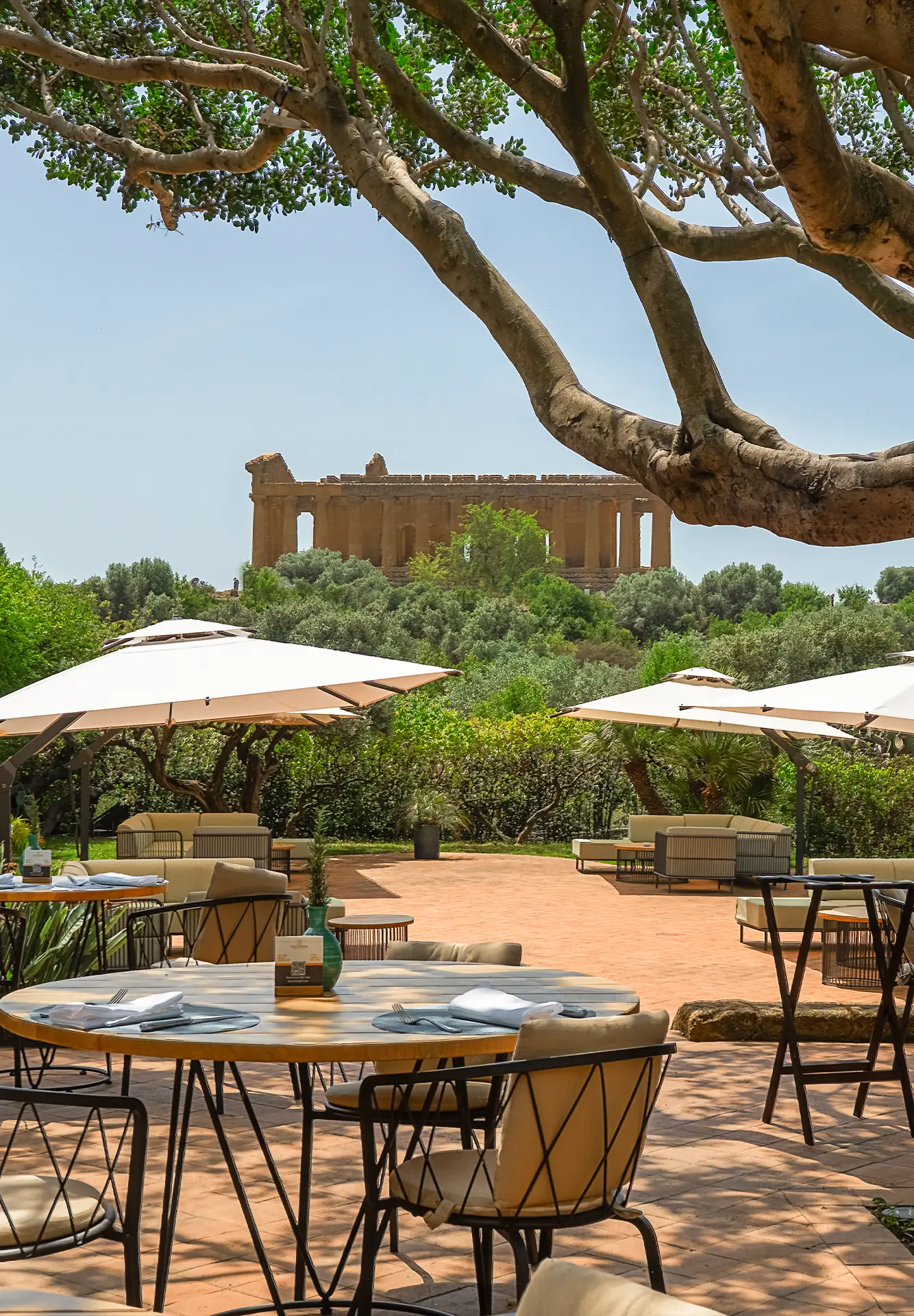 Spazio all'aperto del Ristorante & Bar Federico II con tavoli, ombrelloni, giardino rigoglioso e vista sul tempio della Valle dei Templi.