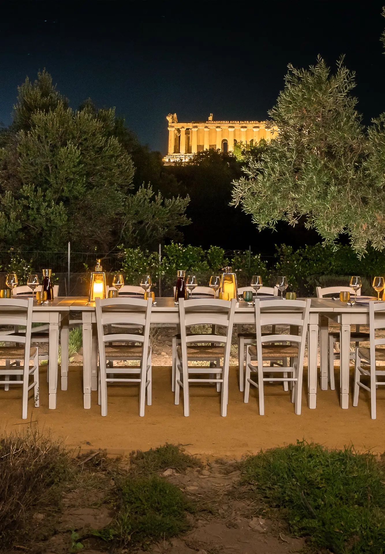 Lunga tavolata all'aperto allestita di sera per l'esperienza Orto Dining, illuminata da lanterne con il Tempio della Concordia illuminato sullo sfondo.